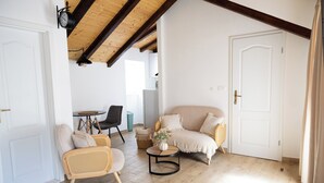 Una stanza con soffitto in legno, pareti bianche, un divano, una sedia, un tavolino con un vaso e una porta.