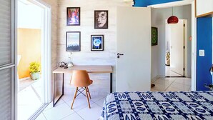 A bedroom with a bed, a desk, and a chair, a door, and a wall with framed pictures.