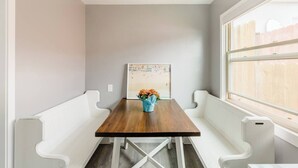 Un coin repas avec une table en bois, des bancs blancs, un tableau encadré au mur et une fenêtre donnant sur un paysage.