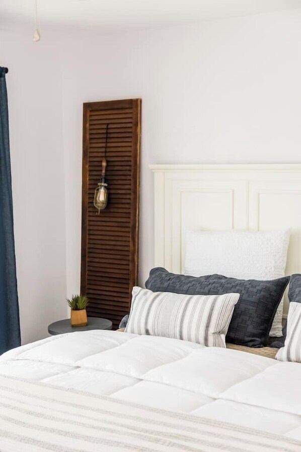 A bedroom with a bed, pillows, a bedside table with a plant, and a wall-mounted lamp. - Cute Beach Cottage - Walk to Beach, Bay, & More (San Diego)