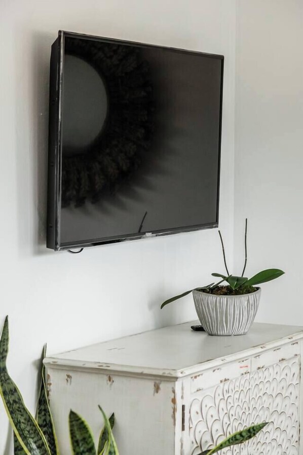 Uma TV de tela plana presa na parede, um armário branco com desenhos decorativos e uma planta em vaso sobre o armário.