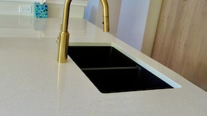 A kitchen with a gold faucet, a white countertop, a black sink, a mirror, and a wooden cabinet.