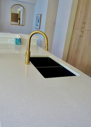 A kitchen with a gold faucet, a white countertop, a black sink, a mirror, and a wooden cabinet.