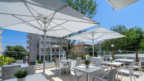 Une terrasse extérieure avec des tables et des chaises blanches, de grands parasols et des plantes en pot.