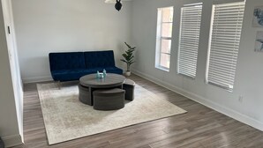A living room with a blue sofa, a round coffee table, a potted plant, and a large window with blinds. - The Burnside Bungalow where Vacation Awaits! (Holiday)
