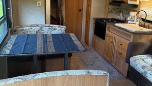 Cocina compacta con microondas, estufa y pileta. Hay una mesa con mantel y un banco. La habitación tiene placares de madera y un ventilador de techo.