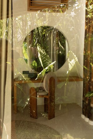 Une pièce dotée d’un grand miroir rond, de meubles en bois et offrant une vue sur la verdure environnante.
