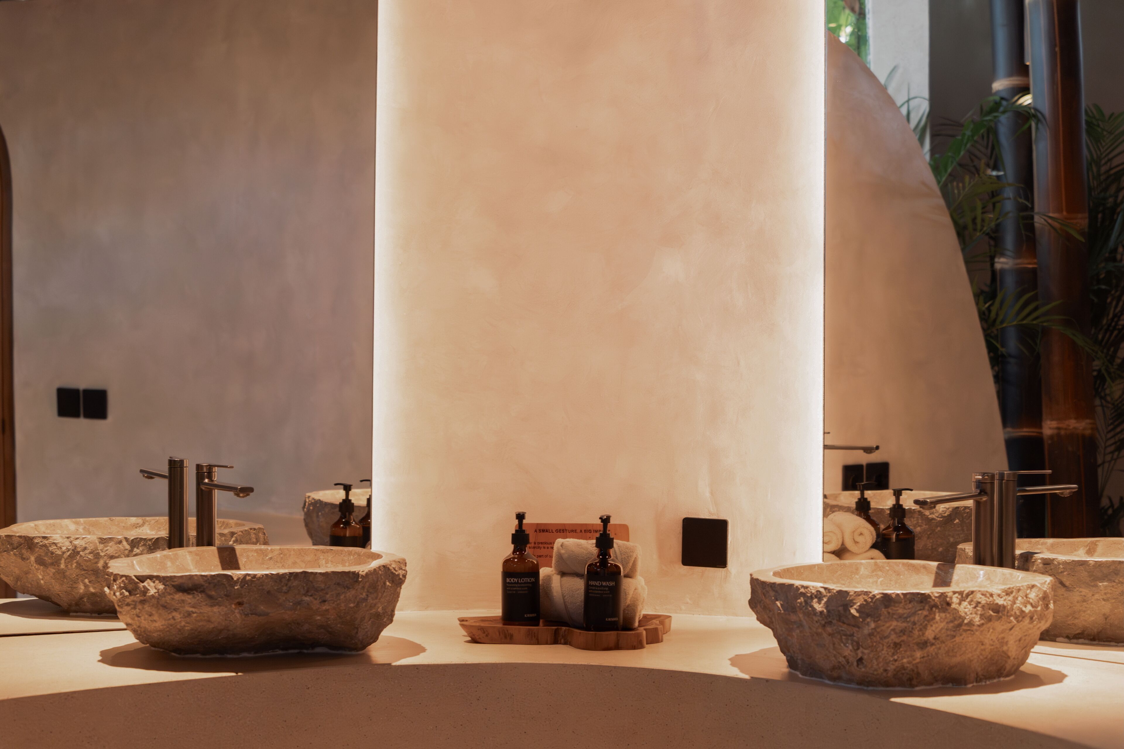 Une salle de bain avec de grands lavabos en pierre, un plateau en bois sur lequel sont posées des bouteilles, et un porte-serviettes.