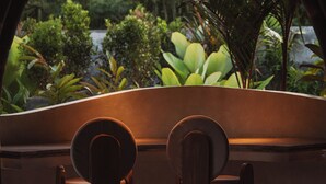 A view through an arched window with a wooden chair in the foreground, leading to a lush garden with various plants and a water feature. - Kaymana (Pecatu)
