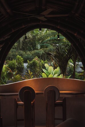 Une vue à travers une fenêtre en arc, avec une chaise en bois au premier plan, qui s’ouvre sur un jardin luxuriant où poussent différentes plantes et où se trouve un élément décoratif aquatique.