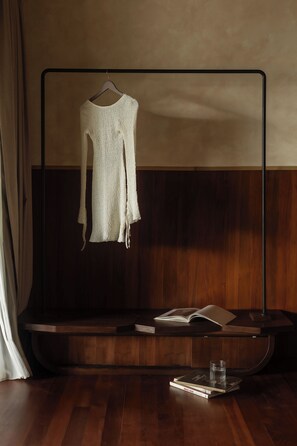 A room with a wooden floor, a white knitted sweater hanging on a metal rack, a book, and a glass on the floor. - Kaymana (Pecatu)