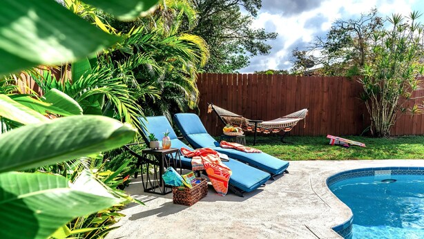Un patio trasero con piscina, sillas de descanso, una hamaca y plantas tropicales.