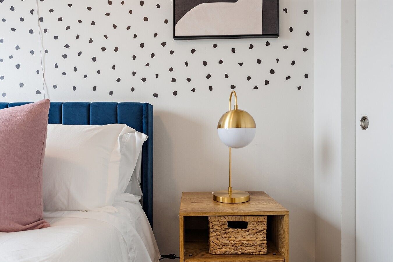 Een slaapkamer met een blauw hoofdbord, witte beddengoed, een roze kussen, een goud- en wit vloerlamp en een klein houten nachtkastje met een gevlochten mandje.