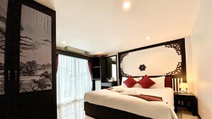 A hotel room with a large bed, a decorative headboard, a nightstand, a wall-mounted artwork, and a window with curtains. - Sunny Beach Hotel (Patong)