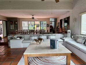 A spacious living area with a sofa, a coffee table, and a dining table. There is a large window providing natural light and a view of the outdoors.