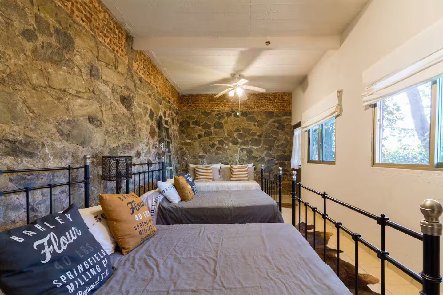 Une chambre avec des murs en pierre, deux lits, un ventilateur de plafond et une fenêtre donnant sur un paysage agréable.