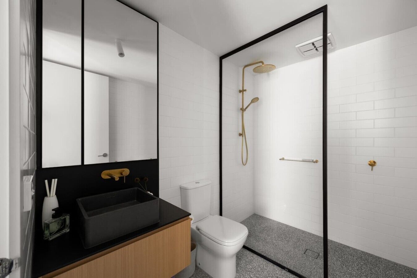 A modern bathroom with a black sink, a toilet, a shower with a gold showerhead, and a large mirror.