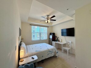 Une chambre à coucher comprenant un lit, un bureau, une chaise, un ventilateur de plafond, une télévision et une fenêtre avec des stores.