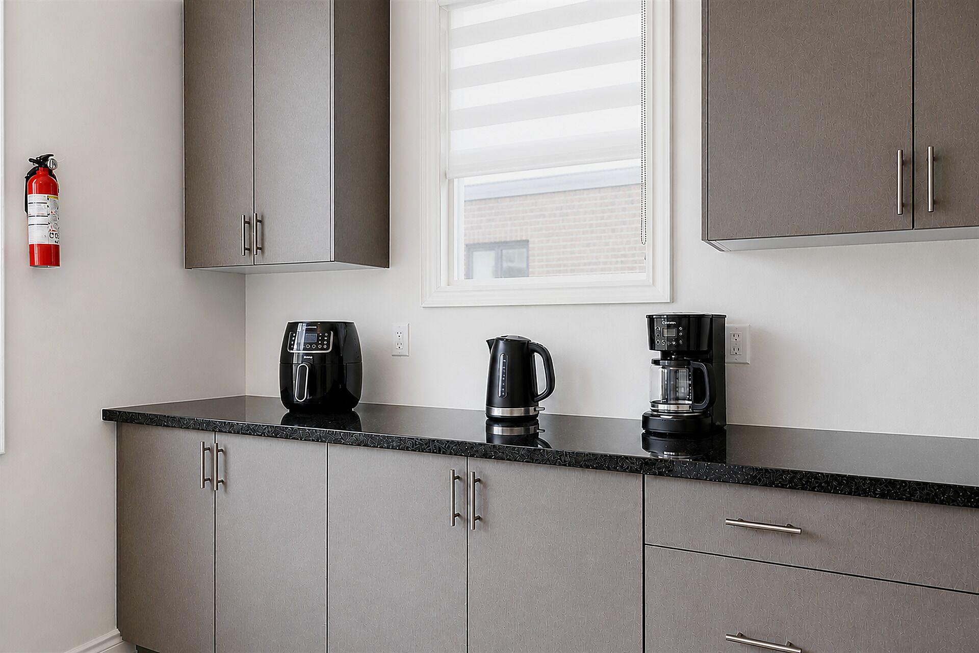 Une cuisine avec des meubles gris, un plan de travail noir, une machine à café, une bouilloire et un extincteur.