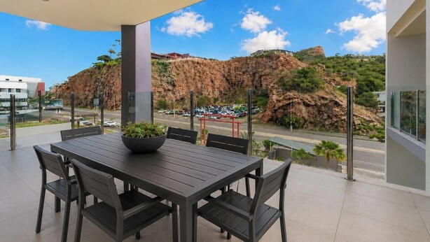 Un espacio moderno para comer al aire libre con una mesa, sillas, una planta en maceta y vistas a una ladera rocosa y un estacionamiento.