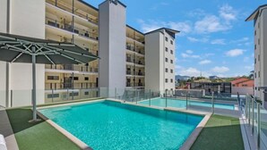 Una piscina con vistas a un edificio de varios pisos, una zona techada con estructura metálica y cielo despejado.