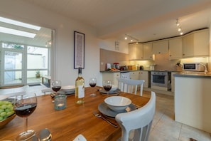 A modern kitchen with white cabinets, a dining table set with wine glasses and a bottle, and a view of the outdoors through large windows. - Little Palm Trees is walking distance of the charming surf-village of Polzeath (Polzeath)