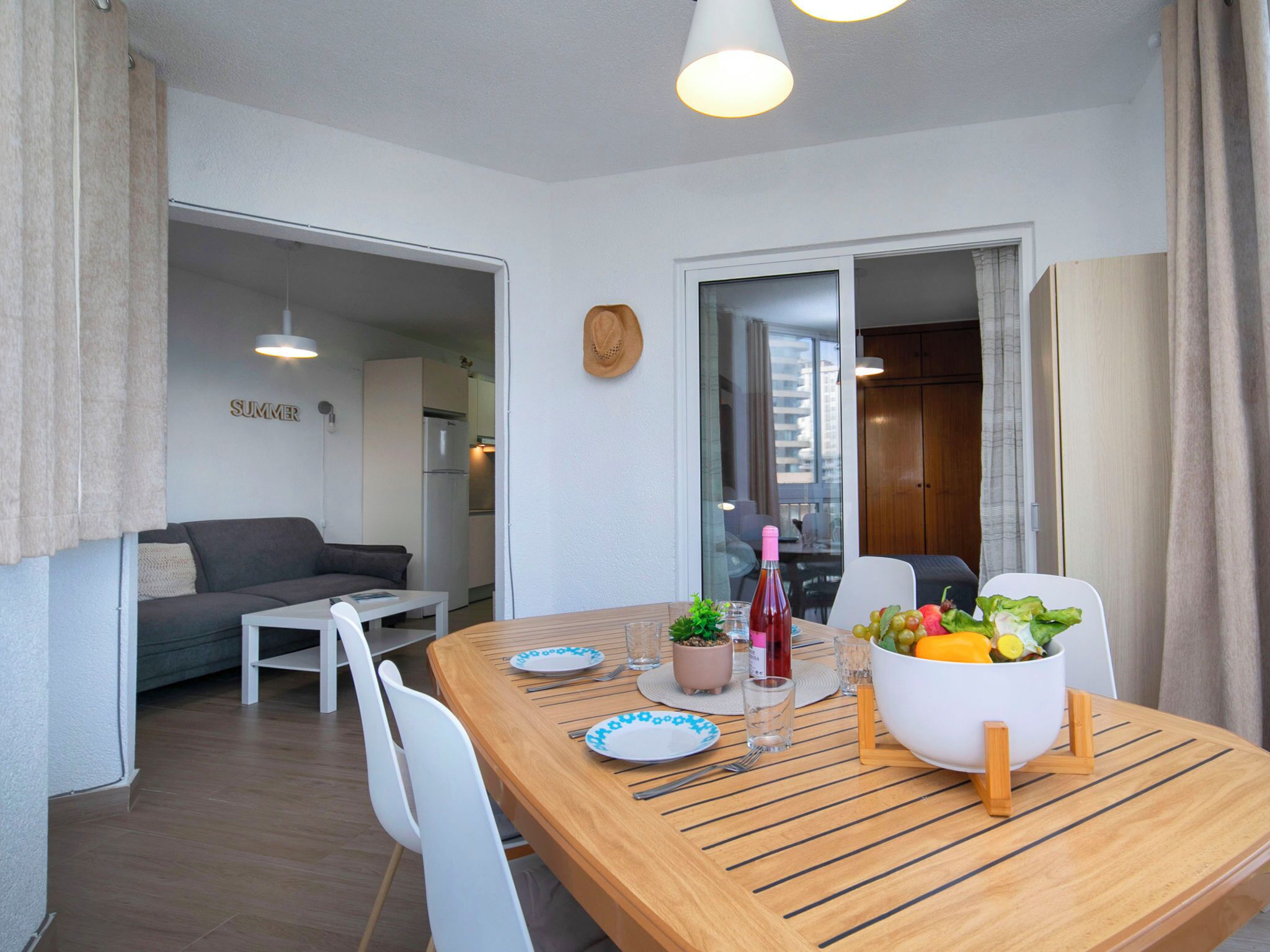 A dining area with a wooden table, white chairs, a bowl of fruit, and a bottle of wine.