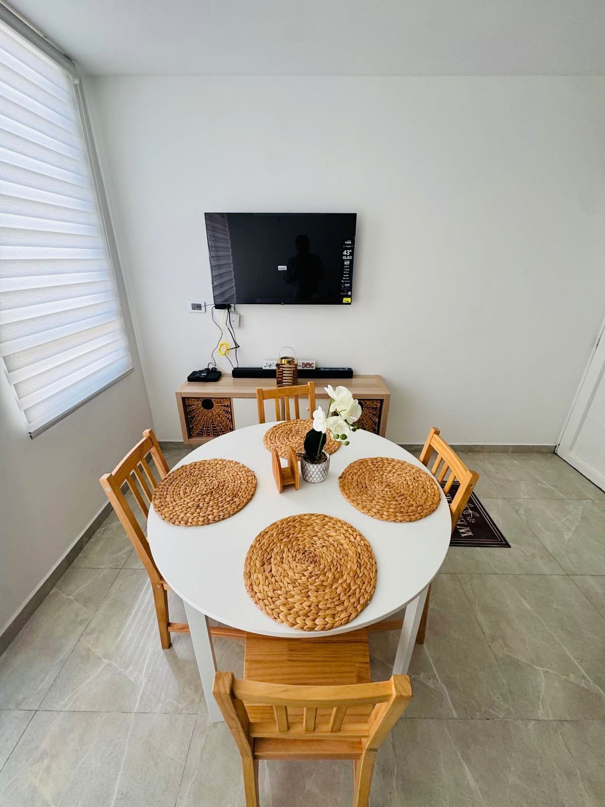 Un coin repas comprenant une table ronde, quatre chaises, un téléviseur fixé au mur et une fenêtre avec des stores.