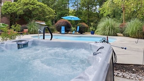 A hot tub with a pool, surrounded by greenery and a patio area with chairs and an umbrella.