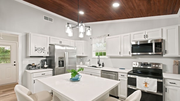 A modern kitchen with white cabinetry, stainless steel appliances, a central island with chairs, and a wooden ceiling.