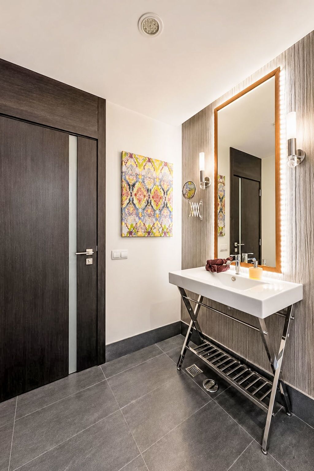 A modern bathroom with a dark wood door, a white vanity with a large mirror, a colorful artwork on the wall, and a light fixture above the mirror.