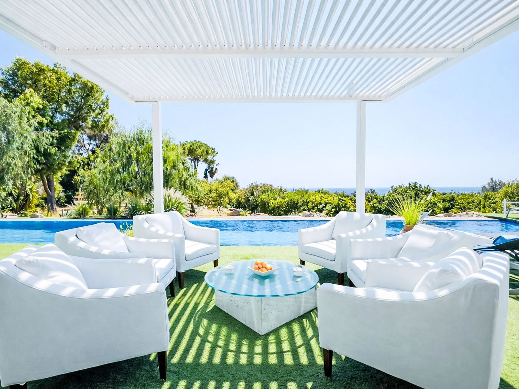 A covered outdoor seating area with white furniture, a glass table, and a pool with a view of the sea.