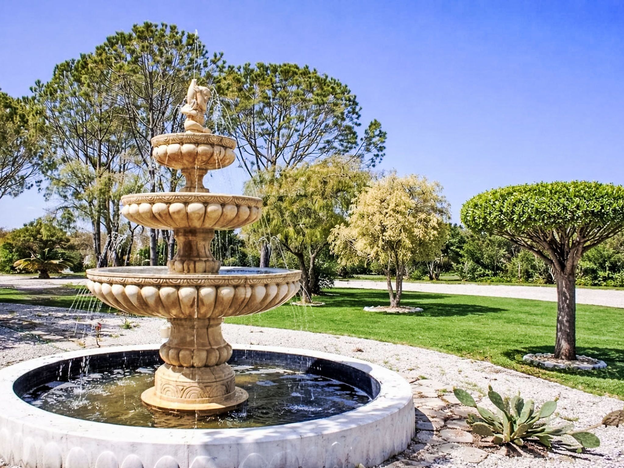 A multi-tiered stone fountain with water flowing, surrounded by a well-maintained garden with various trees and shrubs.