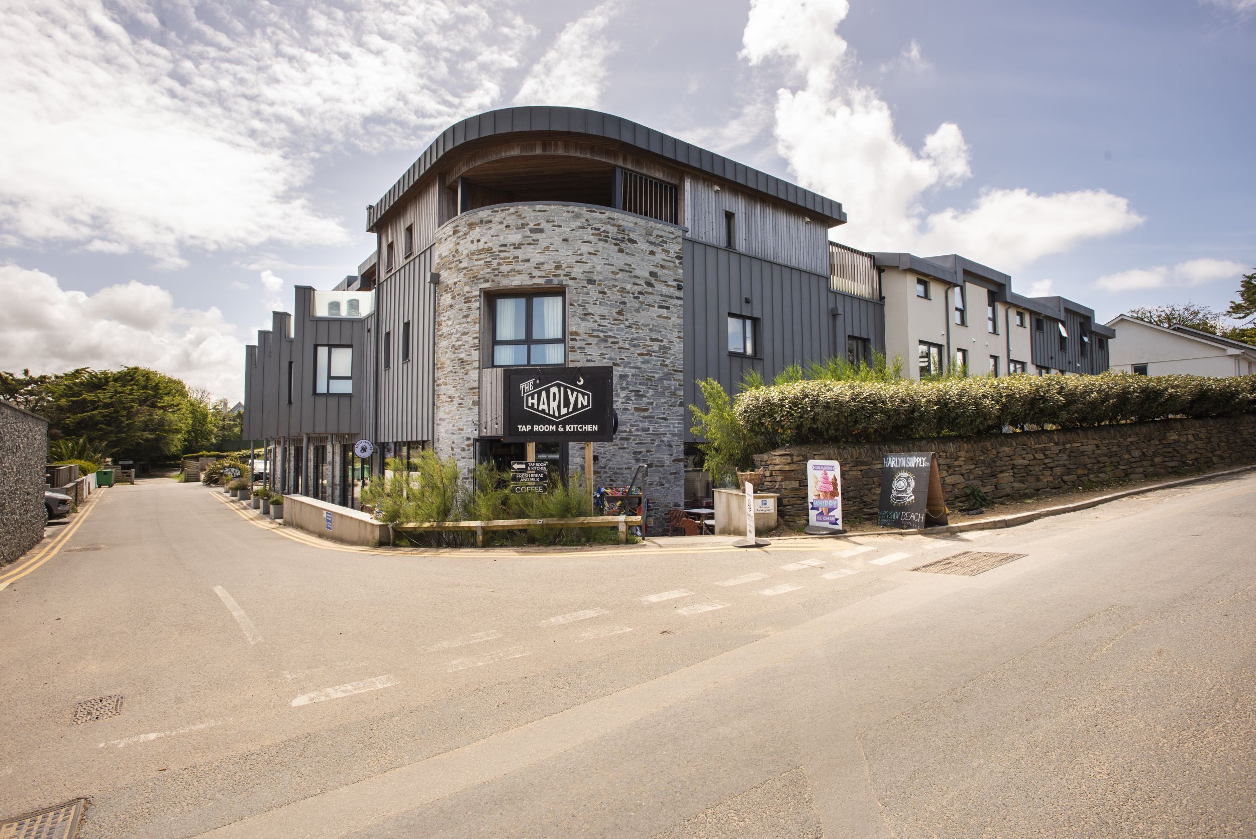 Een modern gebouw met een stenen gevel, een bord met 'The Harlyn Tap Room & Kitchen' en een klein terras.