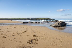 A beach with footprints, a rocky outcrop, and a distant view of a hill with buildings. - Treth Nook - Dog friendly, 100m from beach, Parking (Harlyn Bay)