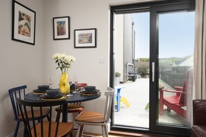 A dining area with a table set for two, chairs, a vase with flowers, and framed pictures on the wall. - Treth Nook - Dog friendly, 100m from beach, Parking (Harlyn Bay)