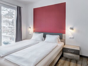 A hotel room with a bed, two bedside tables, a red accent wall, and a window with a view of a snowy landscape. - LoriVita by Interhome (Saalbach-Hinterglemm)