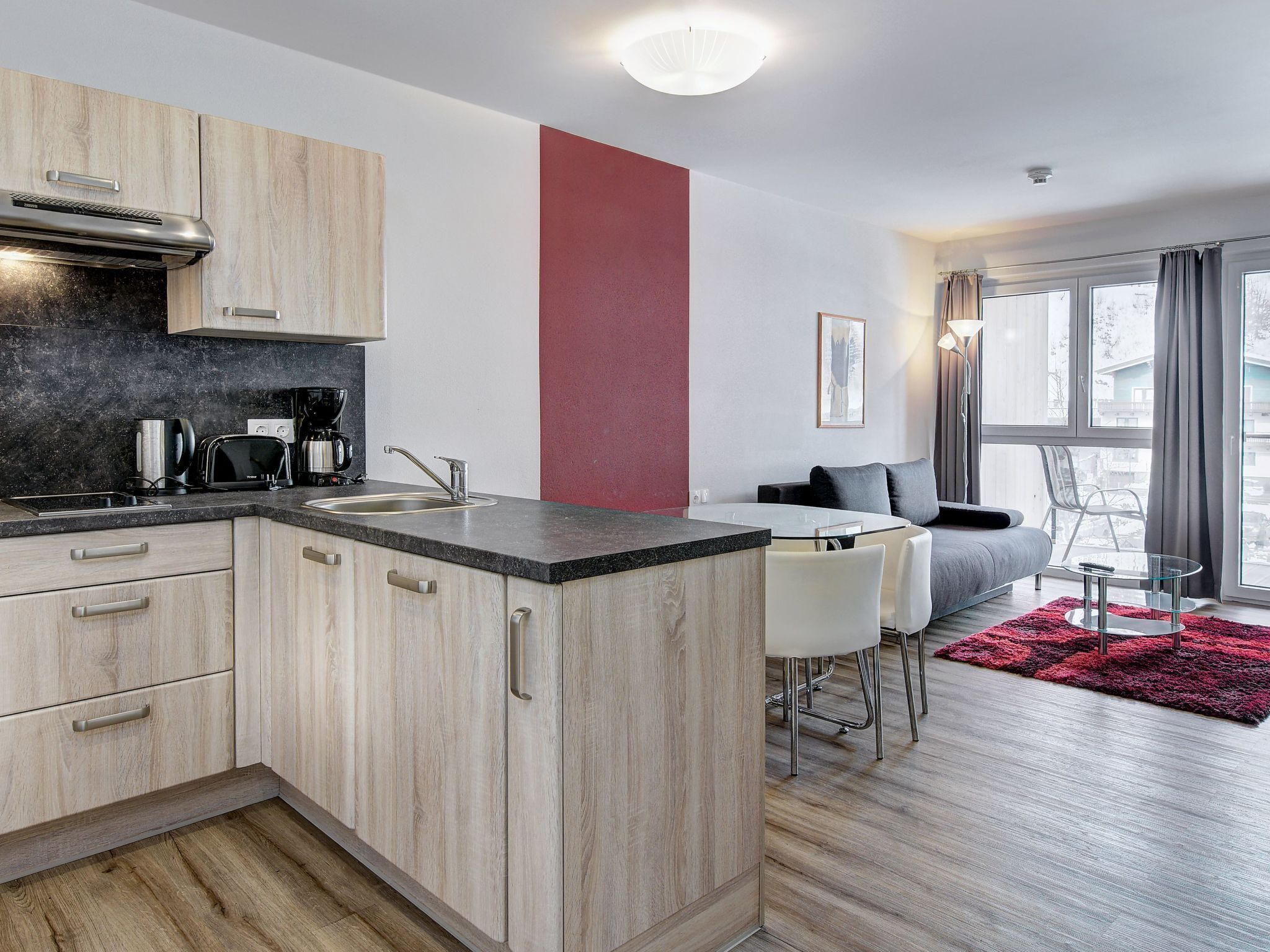 A modern kitchen with wooden cabinets, a black countertop, and a coffee maker. There is a dining area with a glass table and chairs, and a red rug.