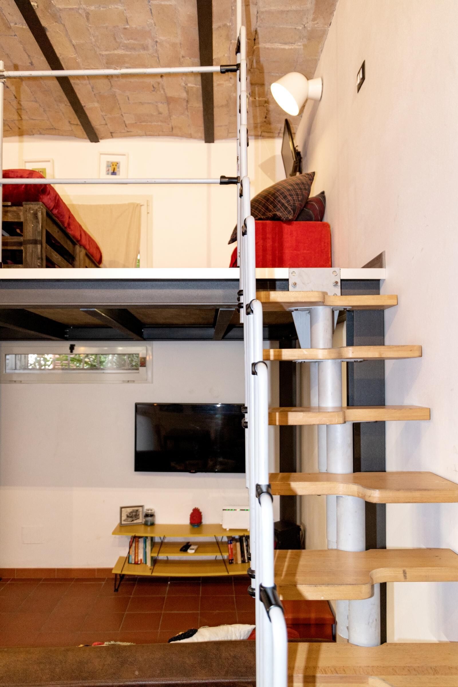 Ein kompakter Wohnraum mit Schlafkoje, einer Treppe, einem Fernseher und einem kleinen Tisch mit Büchern und einer Lampe.