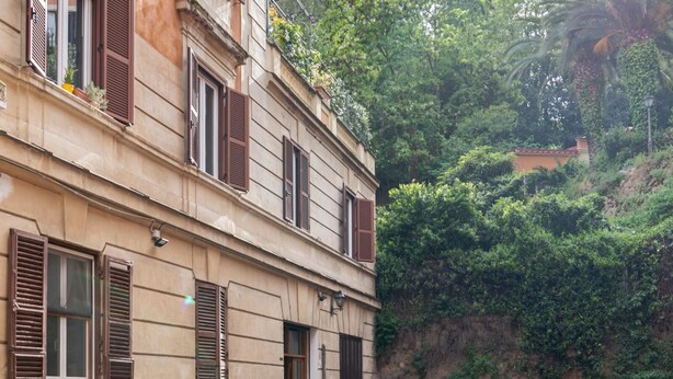 A multi-story building with arched windows and shutters, a paved courtyard, and lush greenery in the background. - Trastevere Secret Loft (ROMA)