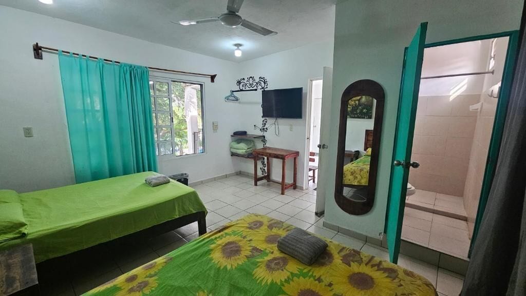 Quarto com duas camas, uma escrivaninha, televisão, espelho e área de chuveiro.