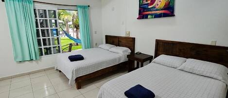 Habitación de hotel con dos camas, una ventana con cortinas, una hamaca afuera y un cuadro colorido en la pared.