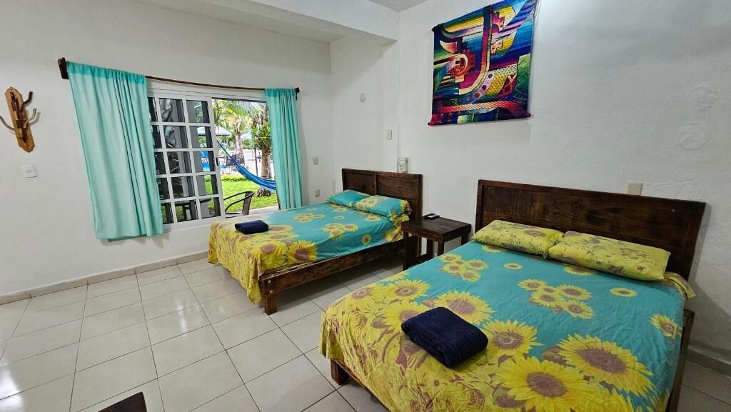 Una habitación con dos camas, un ventanal con cortinas, un tapiz de colores y piso de baldosas.