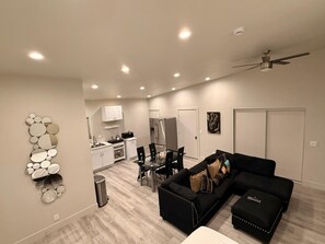 Ein modernes Wohnzimmer mit einem schwarzen Ecksofa, einer Essecke mit Tisch und Stühlen, einer Küche mit weißen Schränken und einem Deckenventilator.