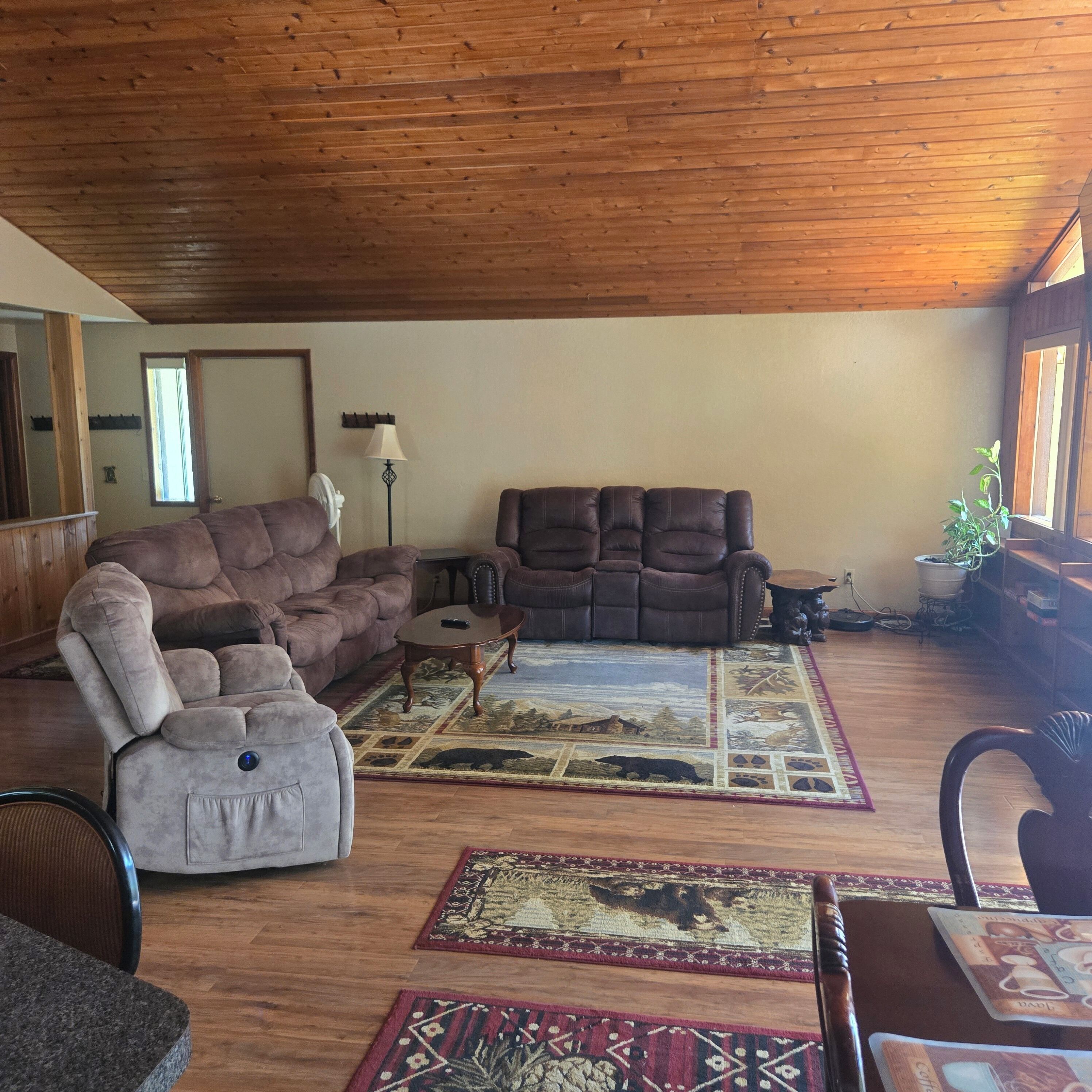 A spacious living room with a high wooden ceiling, a brown leather sofa, a grey armchair, and a coffee table.