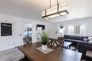 A modern living room with a wooden dining table, a sofa, and a large window with a view.