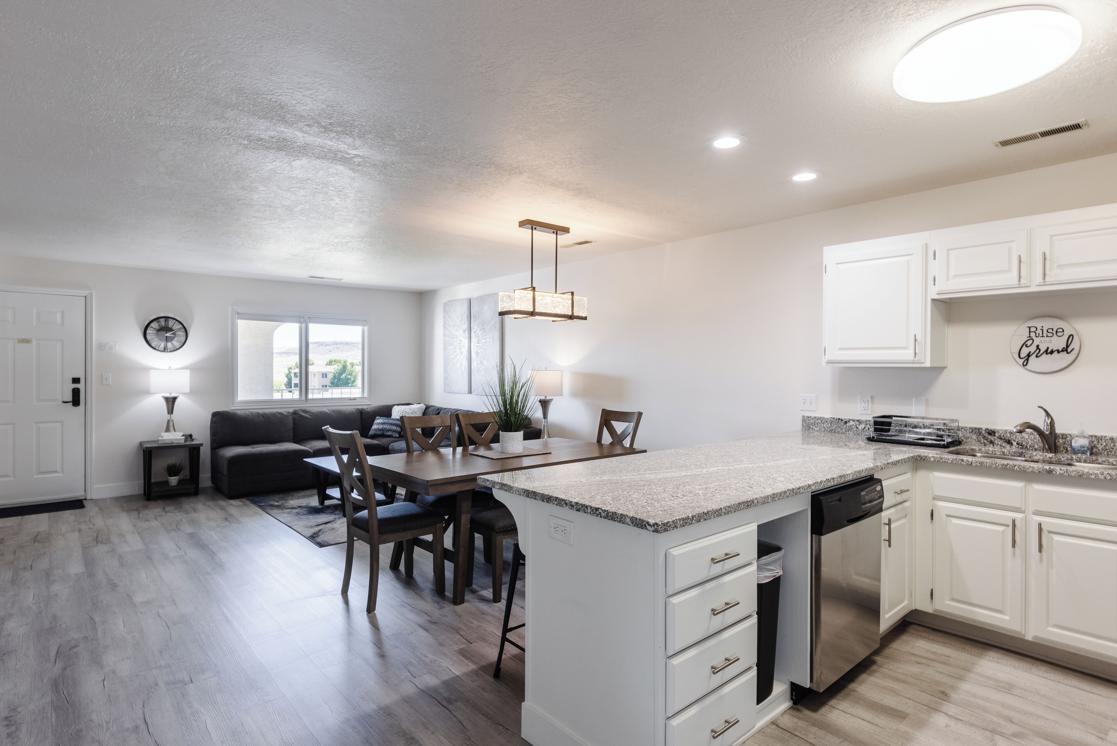 A modern kitchen with white cabinets, a granite countertop, and a dishwasher. There is a dining area with a table and chairs, and a living room with a sofa and a window.