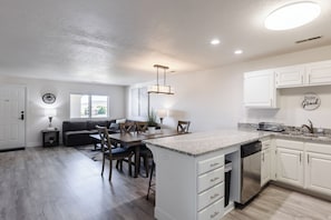 A modern kitchen with white cabinets, a granite countertop, and a dishwasher. There is a dining area with a table and chairs, and a living room with a sofa and a window.