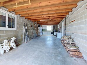 Un grand garage avec un sol en béton, des poutres en bois au plafond, un lave-linge et une pile de bois de chauffage.