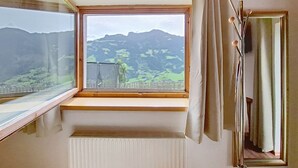 Quarto com uma janela ampla que dá para uma paisagem montanhosa, um radiador abaixo da janela e um espelho com luminária.